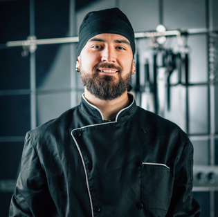 Chef de cozinha com pele caucasiana, barba, sorriso no rosto, usando toca e camisa de chef de cozinha, tudo na cor preta com sua cozinha ao fundo.